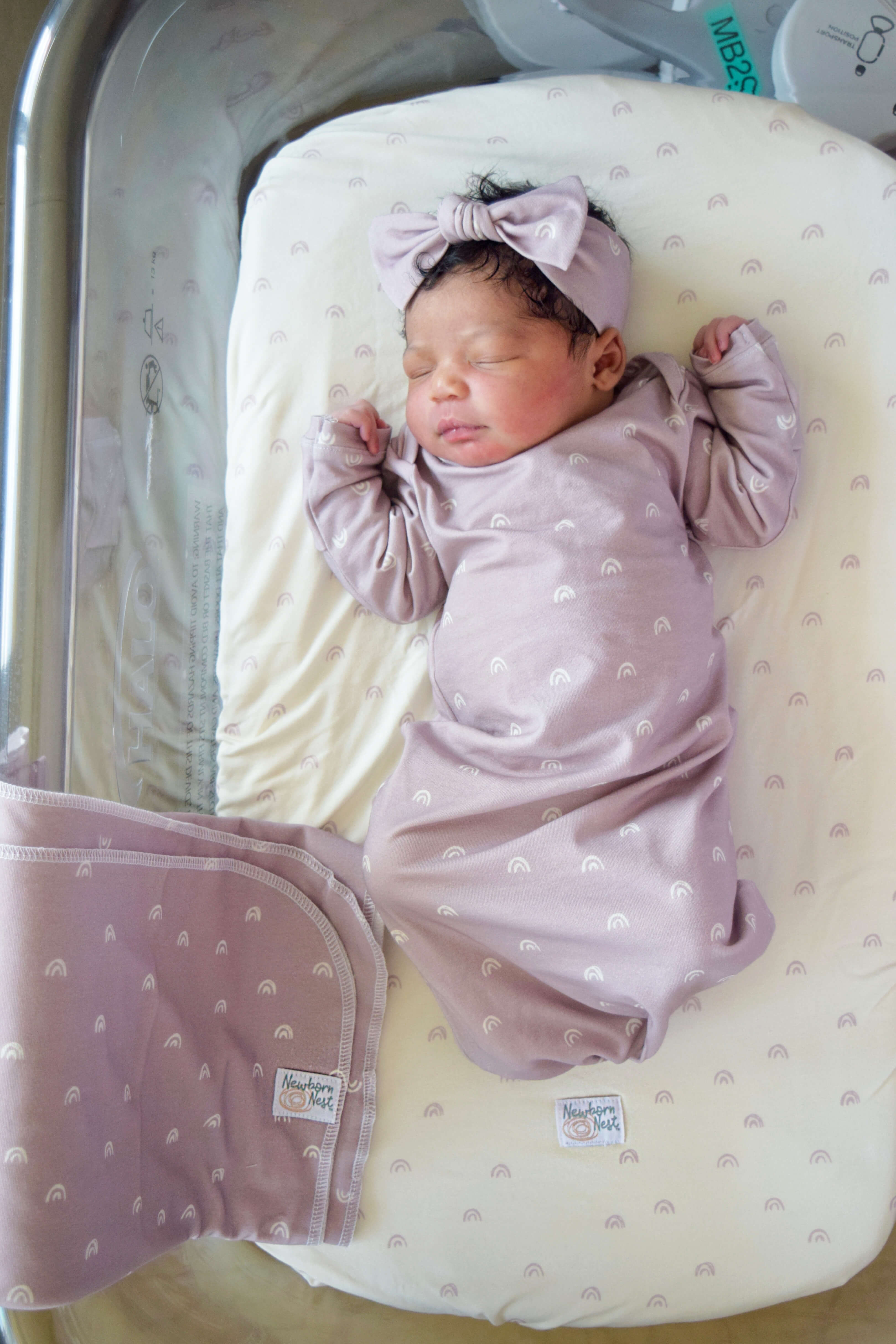African American baby in bassinet. Gown,bow, swaddle are lilac color with ivory rainbows. Sheet is ivory with lilac rainbows. 
